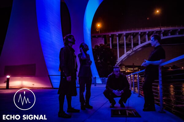 Phaedrus, Charles, John, and Adam (Echo Signal in 2016) under a bridge span at night, lit in magenta and blue, the lit arch of a second bridge behind them.